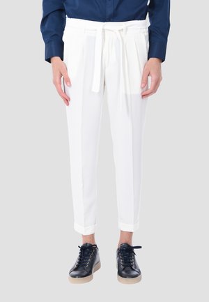 Donna che indossa pantaloni bianchi a vita alta con cintura annodata, camicia blu navy e sneakers nere, in piedi davanti a uno sfondo chiaro.