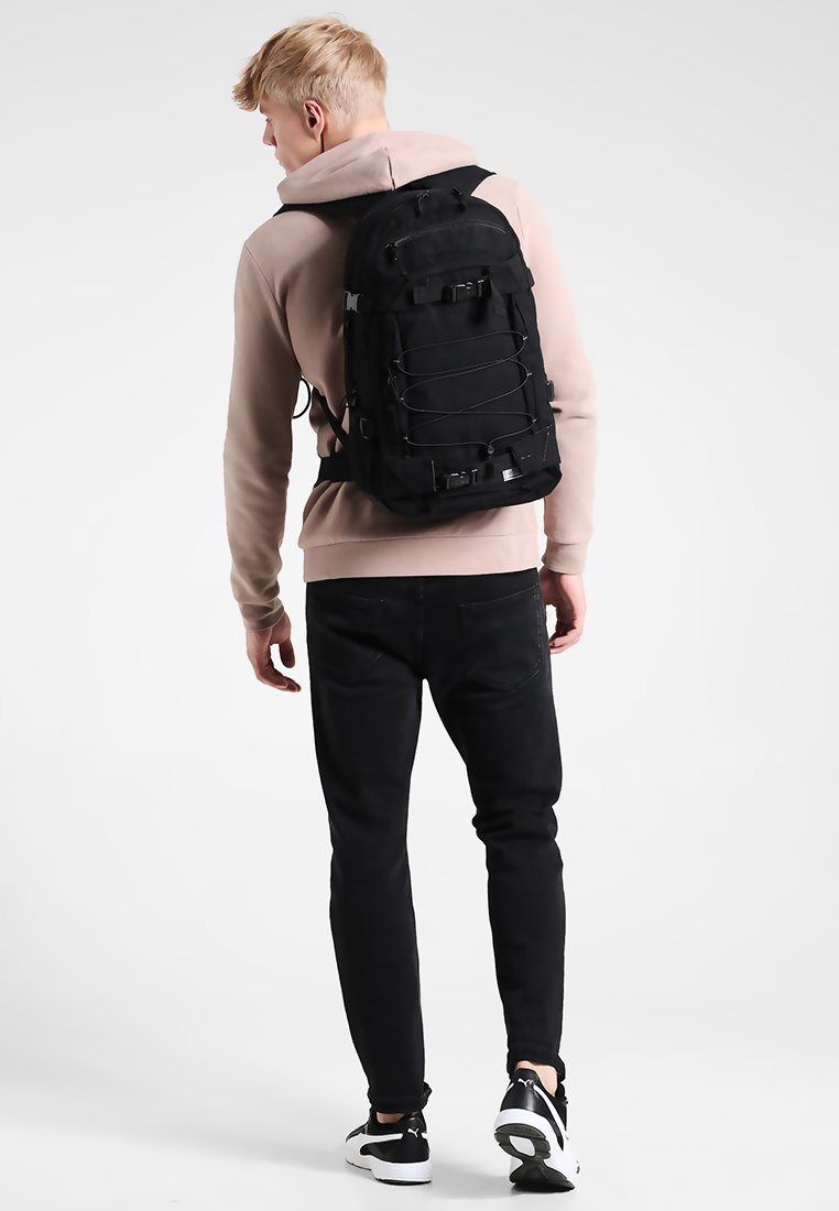 Mochila negra con múltiples correas ajustables, con un exterior de nailon texturizado y una forma redondeada. Se lleva en la espalda, combinada con pantalones negros.