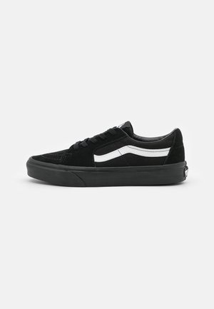 Schwarzer Sneaker mit niedrigem Schaft, weißem seitlichem Streifen, schwarzen Schnürsenkeln und dicker schwarzer Sohle auf hellem, einfarbigem Hintergrund.