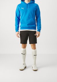 Blå huvtröja med ficka fram, svarta shorts, vita strumpor med svart logotyp och vita träningsskor med bruna sulor.