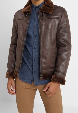 Mężczyzna w brązowej skórzanej kurtce z kożuchem, nałożonej na niebieską koszulę w wzory zapinaną na guziki oraz beżowe spodnie, stojący na jednolitym tle.