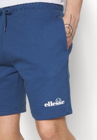 Modré šortky z bavlnenej zmesi s elastickým pásom, bočnými vreckami a bielym logom potlačeným blízko lemu. Mäkká textúra, neformálny dizajn.