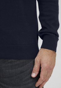 Marineblå strikket genser med ribbestrikkede mansjetter og nederkant. Stoffet virker glatt og har mellomvekt, med fine teksturdetaljer synlige.