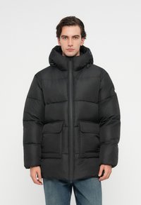 Fekete puffajka kapucnival, elülső cipzárral, két nagy fedeles zsebbel és steppelt mintával. Közepes hosszúságú és laza fazonú.