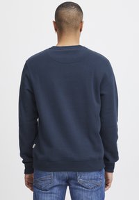 Tumši zila sporta jaka ar apaļu kaklu un garām piedurknēm, izgatavota no mīksta auduma, ar ribotām piedurknēm un apakšmalu, valkāta kopā ar ziliem džinsiem.