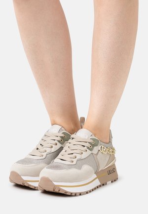 Persoon draagt beige en lichtgrijze platform sneakers met gouden kettingdetail en "LIU·JO" merknaam op de zool tegen een witte achtergrond.