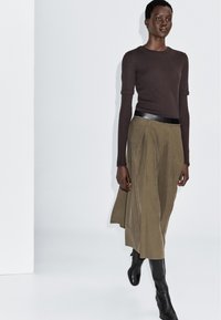 Mannequin portant un haut marron à manches longues, une jupe plissée vert olive avec ceinture noire, et des bottes noires montantes, marchant devant un fond blanc uni.