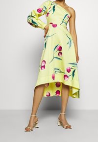 Žena v žlutých květovaných šatech na jedno rameno s dlouhým volným rukávem a asymetrickým lemem, doplněná bílými lodičkami na blokovém podpatku s kotníkovým páskem.