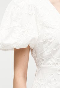 Λευκό φόρεμα με φουσκωτά μανίκια και υφή λουλουδένιας κεντητικής, εφαρμοστό στη μέση, απεικονίζεται από τον ώμο μέχρι τη μέση στο χέρι ενός ατόμου.