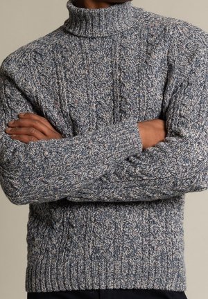 Person trägt einen dicken, grauen Rollkragenpullover aus Kabeleinstrick mit verschränkten Armen, der das strukturierte Strickmuster zeigt.