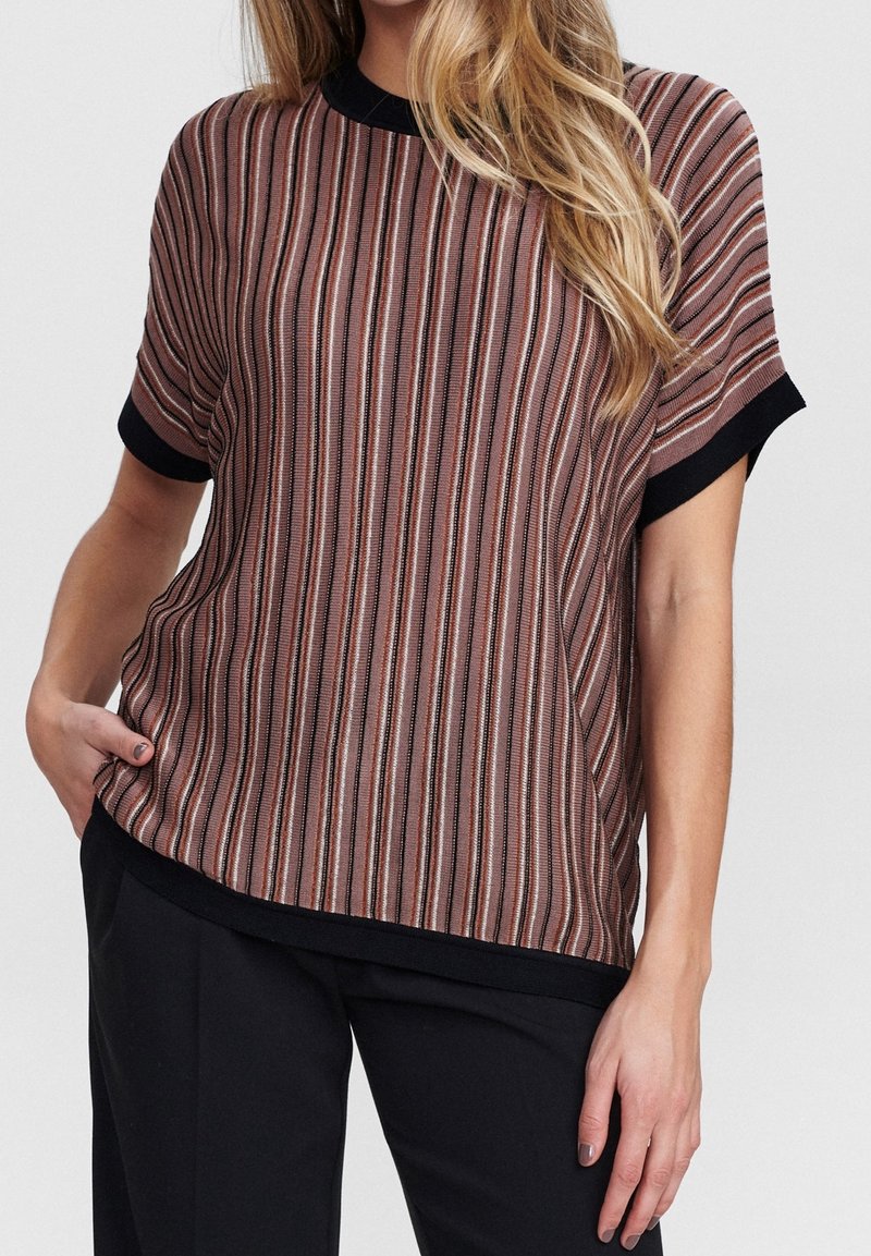 Femme portant un haut à manches courtes rayé marron avec des bordures noires et un pantalon noir, debout avec une main dans la poche.