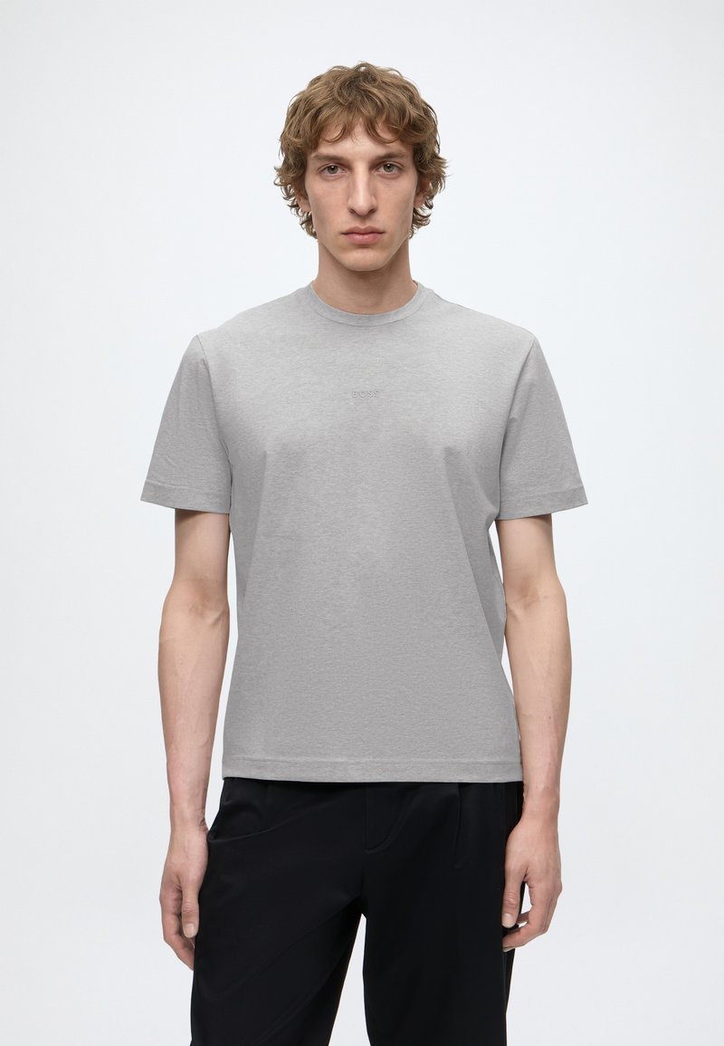 Jeune homme aux cheveux bouclés portant un t-shirt gris clair uni et un pantalon noir, debout devant un fond clair uni.