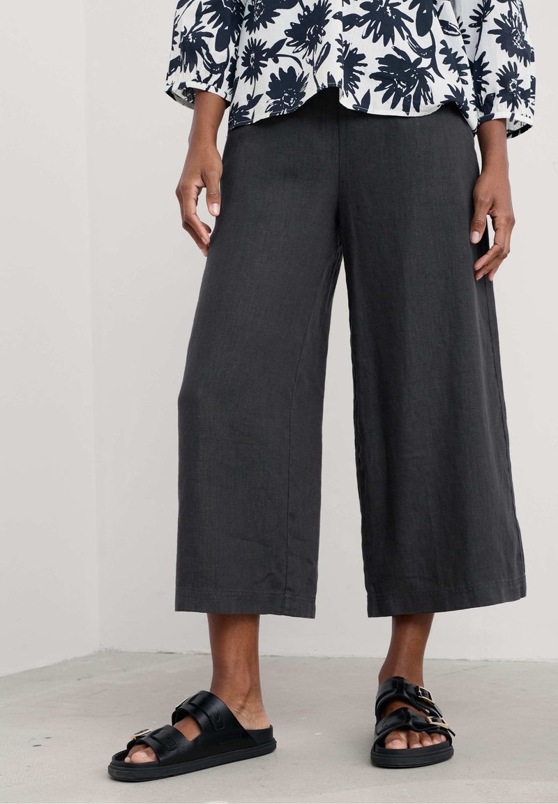 Zwarte wijde linnen broek met een textuurafwerking, gecombineerd met zwarte sandalen met twee banden en een metallic gespdetail.