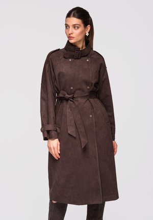 Frau trägt einen dunkelbraunen Trenchcoat mit Gürtel und doppelreihigen Knöpfen, steht und schaut seitlich vor einem einfarbigen Hintergrund.