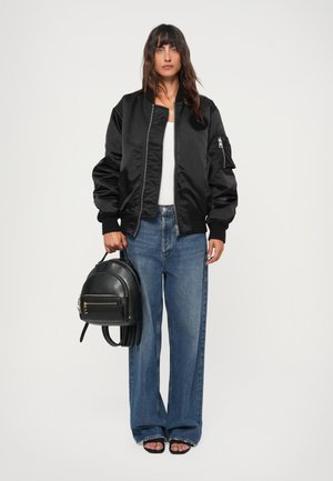 Svart bomberjakke med ribbekant, kombinert med høy midje blå jeans og svart ryggsekk. Modellen står med svarte åpen tå sandaler.