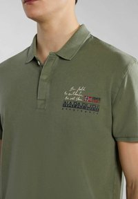 Polo verde realizzato in tessuto texturizzato, con colletto, patta con bottoni e testo stampato con marchio ed emblema della bandiera.
