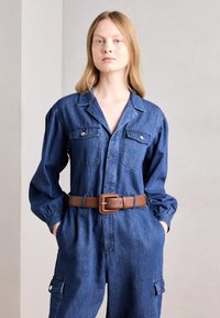 Farmer overál hosszú ujjakkal, két mellzsebbel, barna bőövvel ellátott derékkal és laza szabással.