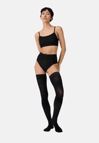 Schwarze Strumpfhosen, hochgeschnittene Unterwäsche und ein kurzes Bustier mit dünnen Trägern. Das Material ist glatt mit matter Oberfläche und minimalen Markenakzenten.