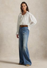Femme portant une blouse paysanne blanche à manches longues, jean bleu à jambes larges avec poches avant, ceinture marron et chaussures beiges, debout devant un fond uni.