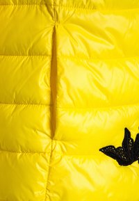 Gul vadderad väst gjord av glansigt syntetmaterial. Har en kviltad design, vertikal slits och en svart broderad fjäril som detalj.