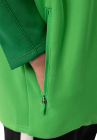 Grüner Sportshirt-Stoff mit glatter Textur, ausgestattet mit einer seitlichen Reißverschlusstasche und kontrastierenden Nähten entlang der Ärmelkanten.