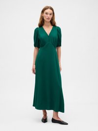 Woman stands wearing a dark green midi dress with puffed short sleeves and black pointed flats against a plain white background.