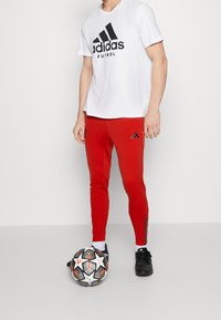 Bílý tričko Adidas s černým logem, v kombinaci s červenými slim kalhotami se bočními pruhy. Černé fotbalové boty a vzorovaný míč na zemi.