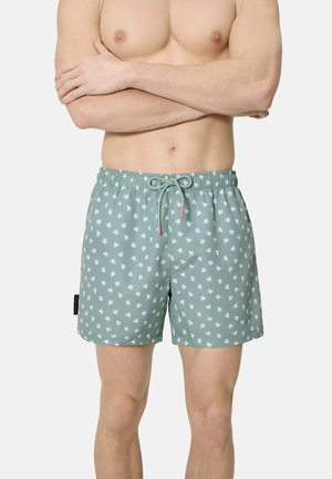 Hombre con pantalones cortos de baño verde claro con pequeños patrones blancos de pulpos, de pie con los brazos cruzados sobre el pecho descubierto.