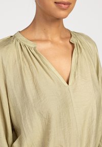Hellgrüne Bluse mit V-Ausschnitt, gefälteltem Halsbereich und weichem, strukturiertem Stoff. Lockere Passform mit unsichtbaren Nähten.