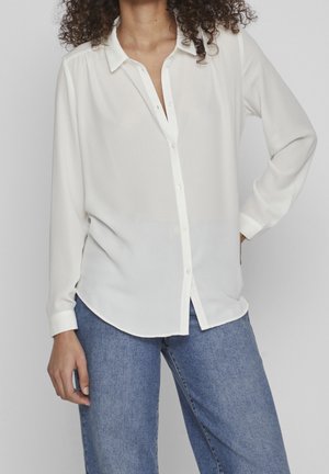 Vrouw draagt een wit bloesje met lange mouwen en knopen, en een blauwe jeans, staand met één hand op de heup tegen een effen achtergrond.