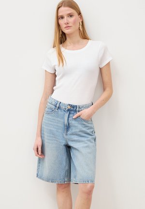 Frau mit langen blonden Haaren, die goldene Ohrringe, ein weißes kurzärmeliges Oberteil und hellblaue, knielange Jeansshorts trägt und vor einer weißen Wand steht.