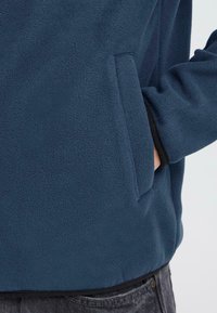 Tmavomodrý fleece pulóver s jemnou textúrou, bočnou vreckom a uvoľneným strihom so zakončením v páse.