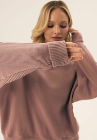 Sweter z dzianiny waffle, z długim rękawem w kolorze mauve, charakteryzujący się luźnym krojem i prążkowanymi mankietami. Materiał jest miękki i lekko elastyczny.