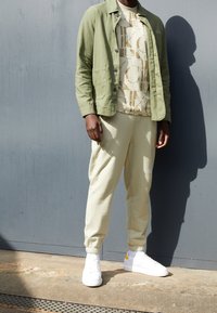 Ljus olivgrön jacka över en beige t-skjorta med grafiskt tryck, i kombination med ljusbeiga sweatpants och vita sneakers med gula accenter.