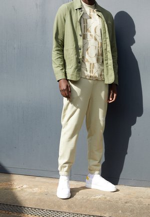 Licht olijfgroene jas over een beige graphic t-shirt, gecombineerd met lichtbeige sweatpants en witte sneakers met gele accenten.