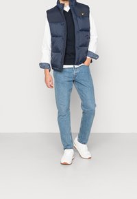 Chaleco acolchado azul marino sobre un suéter negro y una camisa blanca, combinado con jeans azul claro y zapatillas blancas con suelas marrones.