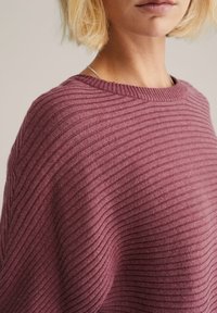 Pletený svetr v tmavě růžové barvě, s horizontální žebrovanou texturou, širokým výstřihem a volným střihem, zdůrazňující měkkost a teplo.