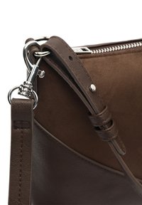 Gros plan sur une bandoulière en cuir brun foncé attachée à un fermoir en métal argenté et une fermeture éclair d’un sac assorti en suède marron.