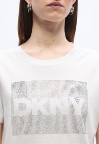 Γυναίκα με λευκό μπλουζάκι με το ασημένιο λογότυπο DKNY με τρουκς και ασημένια κρίκους σκουλαρίκια, με φόντο μια απλή λευκή επιφάνεια.