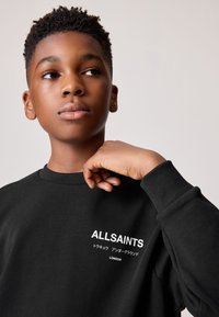 Teenager s krátkými kudrnatými vlasy, který nosí černou mikinu s bílým nápisem "ALLSAINTS LONDON", dívá se bokem s rukou u ramene.