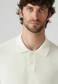 Vit polotröja med en strukturerad väv, med en klassisk krage och tre knappar. Inga synliga mönster eller detaljer.