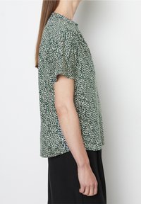 Blusa verde a maniche corte con un motivo maculato bianco. Materiale leggero, scollatura rotonda e vestibilità rilassata. Venti laterali all'orlo.