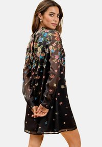Rochie florală neagră cu mâneci lungi din material transparent, având un model cu flori multicolore. Detaliu în formă de cheie la spate, croi lejer și luciu al materialului.