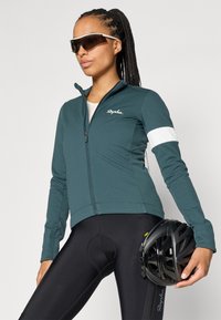 Chaqueta de ciclismo color verde azulado con un alto cuello y una franja blanca. Ajustada, de mangas largas, combinada con pantalones cortos de ciclismo acolchados negros y un casco negro.