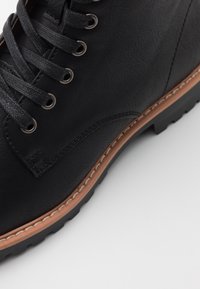 Bottine en cuir noir avec œillets en métal, surface texturée, bout rond et semelle marron contrastante, présentant un détail de couture le long du bord.