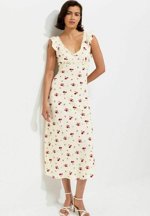 Mujer con vestido midi crema sin mangas con estampado floral rojo y tirantes con volantes, combinado con tacones blancos con tiras, de pie contra una pared blanca.