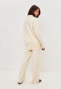 Pulover supradimensionat de culoare crem, cu guler jos, asortat cu pantaloni largi de aceeași nuanță. Textură moale, cusături minime și croială relaxată.