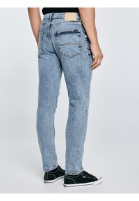 Mees, kes kannab hele siniseid kitsaid teksapükse ja musti madalaid tenniseid, seisab seljaga vaataja poole täiesti tavalise tausta ees.