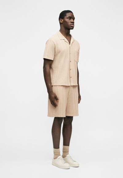 Homme debout portant une chemise beige à manches courtes texturée avec boutons et un short assorti, chaussettes beiges et baskets blanches sur fond blanc.