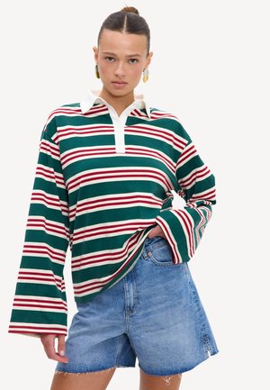 Femme avec les cheveux en chignon, portant de grandes boucles d'oreilles, un t-shirt à manches longues rayé vert et rouge avec col blanc, et un short en jean bleu.